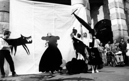 Przestrzeń miejska. Pod białą płachtą stoi pięcioosobowa grupa aktorów i aktorek. Wszyscy trzymają w rękach dwuwymiarowe czarne rekwizyty wyglądające na wycięte z papieru. Aktor po lewej stronie trzyma wilka. Aktorka w centrum przymierza sukienkę. Dwóch aktorów i jedna aktorka po prawej stronie trzyma takie rekwizyty, jak piła do cięcia drewna, domek i ryba. Po prawej stronie stoją widzowie oglądający aktorów. Za ich plecami stoi budynek.