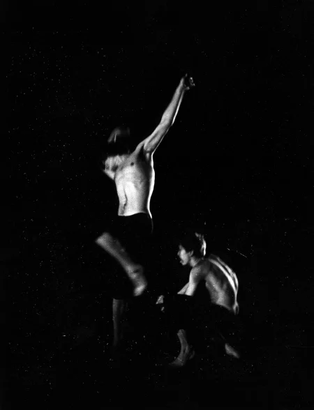 W centrum aktor uchwycony w skoku. Ma ręce wyciągnięte do góry. Ubrany jest w czarne spodenki, ma nagi tors. Nie widać jego głowy. Po jego prawej stronie klęczy aktor. Jest ubrany w czarne spodenki, ma nagi tors.