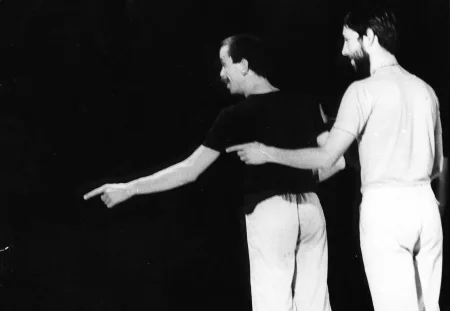 Dwóch aktorów stojących plecami w stronę widza. Mają głowy zwrócone w bok. Uśmiechają się i wskazują coś palcem. Obaj ubrani są w jasne spodnie. Jeden ubrany jest w czarną koszulkę, drugi w białą.