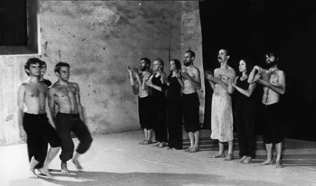 Grupa aktorów i aktorek stojących w szeregu na tle czarnej płachty. Aktorzy ubrani są w czarne spodenki, mają nagie torsy. Aktorki ubrane są w czarne spodnie i czarne koszulki. Wśród grupy wyróżnia się aktor ubrany w jasne spodenki. Wszyscy są boso. Po lewej stronie na ugiętych nogach przechodzi dwóch aktorów. Za nimi idzie słabo widoczna aktorka.