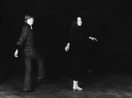 Na scenie stoją zwrócone twarzą do widza dwie aktorki. Przyjmują identyczną pozę. Stoją na lewej nodze, podczas gdy prawa jest lekko uniesiona. Mają lekko uniesione ręce i skręcają tułów. Aktorka po lewej stronie ubrana jest w czarne buty, spodnie jeansowe i sweter w paski. Aktorka po prawej stronie ubrana jest w biały buty i czarną sukienkę.