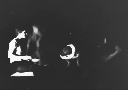 Niewyraźne, rozmazane zdjęcie. Po lewej stronie klęczy aktor zwrócony profilem do widza. W centrum stoi na ugiętych nogach, skulony aktor obejmujący sam siebie rękami. Po jego lewej i prawej stronie niewyraźne sylwetki aktorów.