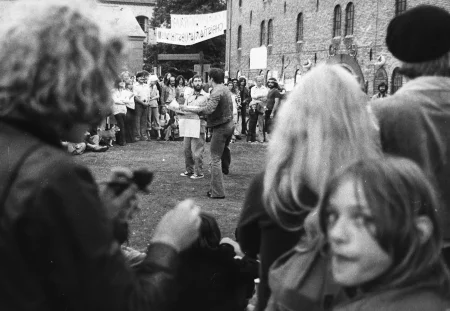 Plener. Na ziemi, w kręgu utworzonym przez widzów stoją dwaj aktorzy. Ubrani są w jeansowe spodnie i jeansowe kurtki. Wyglądają, jakby rozmawiali. Trzymają w rękach mapy albo gazety. Na pierwszym planie w aparat spogląda młoda dziewczyna. W tle widać budynki.