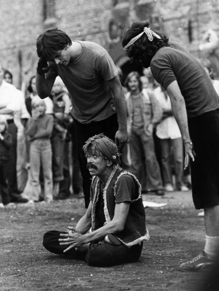 W centrum siedzi aktor w siadzie skrzyżnym. Wygląda, jakby medytował. Widać tylko jego białka oczu. Na głowie ma zawiązaną chustę. Obok niego stoi dwóch aktorów w czarnych spodenkach i podkoszulkach. W tle widać stojących widzów.