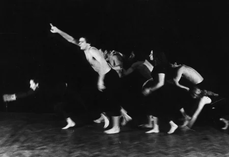 Grupa aktorów i aktorek ekspresyjnie porusza się w lewą stronę. Są boso, mają czarne spodenki, niektórzy mają czarne koszulki, inni są bez koszulek. Wyglądają, jakby rywalizowali w wyścigu. Znajdują się na podłodze z paneli.