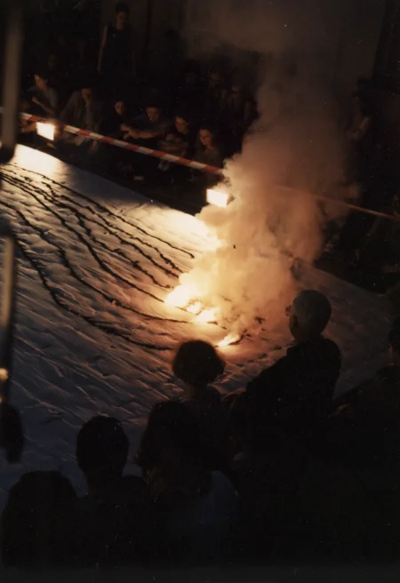 Scena ustawiona w przestrzeni miejskiej. Scena jest biała. Leżą na niej długie wzory w różnych kolorach. W niektórych miejscach są podpalone. Unosi się z nich dym. Pod sceną siedzi i stoi publiczność.