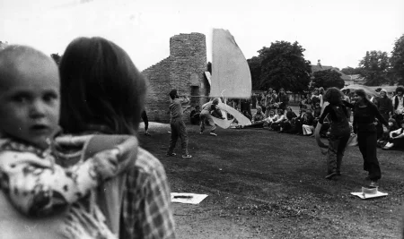 Plener, ruiny jakiegoś budynku. Troje aktorów energicznie macha białymi flagami i wstęgami. W tle siedzą i stoją widzowie, którzy oglądają aktorów. Na pierwszym planie widać plecy widzki trzymającej niemowlę na plecach.