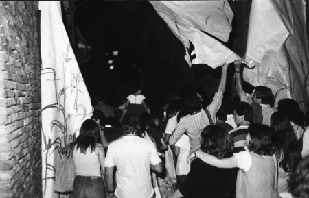 Przestrzeń miejska. Tłum ludzi zwróconych w stronę widza plecami. Idą przed siebie. Niektórzy dotykają strzępy płachty zwisającej z góry.