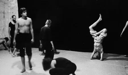 Aktorzy i aktorki chodzą powoli na tle czarnej płachty. Aktorzy ubrani są w czarne spodenki, mają nagie torsy. Aktorki ubrane są w czarne spodnie i czarne koszulki. Pod czarną płachtą stoi na głowie aktor ubrany w jasne spodenki. Jego tors jest nagi.