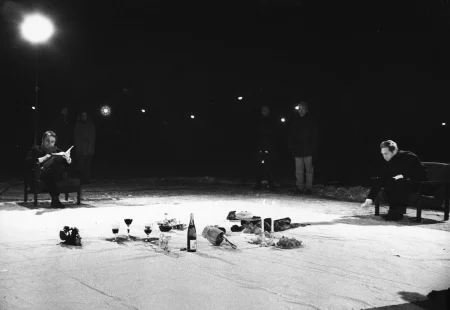 Na białej płachcie leżą przedmioty, takie jak butelki, owoce i kieliszki. Po prawej stronie płachty na krześle siedzi aktor trzymający w ręce napiętą linkę. Z tyłu płachty na krześle siedzi aktor trzymający na kolanach książkę. Aktor mówi coś przez mikrofon. Za jego plecami stoi mała grupa widzów.