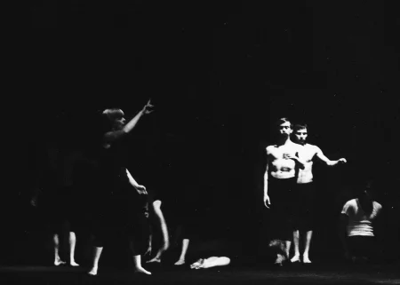 Niewyraźne zdjęcie. Po lewej stronie stoi aktorka ubrana na czarno. Jest boso. Ma jedną rękę wyciągniętą w górę i patrzy w bok. Po prawej stronie, bardzo blisko siebie stoi dwóch aktorów zwróconych twarzą w stronę widza. Mają czarne spodenki i nagie torsy. W tle niewyraźnie widać grupę aktorów i aktorek.