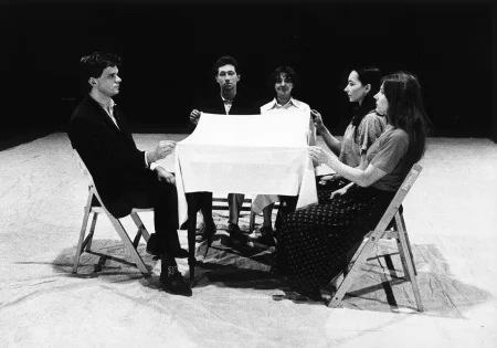 Przy stoliku siedzi pięcioosobowa grupa aktorów i aktorek. Trzymają w rękach biały obrus i unoszą go nad stołem.