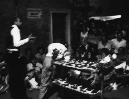 Niewyraźne zdjęcie. Aktor zwrócony plecami do widza przemawia do publiczności. Obok niego stoi kwietnik.