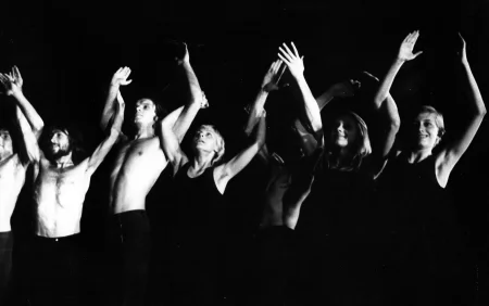 Grupa aktorów i aktorek stojących w szeregu. Aktorzy mają nagie torsy, aktorki ubrane są w czarne koszulki na ramiączka. Patrzą przed siebie, uśmiechają się. Mają ręce uniesiono wysoko w górę.