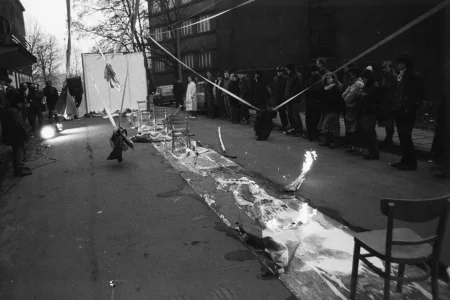 Przestrzeń miejska. Na środku ulicy rozwinięty wybieg. Wybieg jest mokry. Stoją na nim krzesła. W niektórych miejsach wybiegu pali się ogień. Po obu stronach wybiegu stoi publiczność. W tle widać budynki, drzewa i samochody.