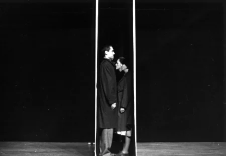 Grupa aktorów i aktorek stoi ściśnięta pomiędzy dwiema ścianami ustawionymi blisko na siebie na środku sceny. Stoją profilem w stronę widza i twarzą do siebie. Mają na sobie czarne płaszcze.