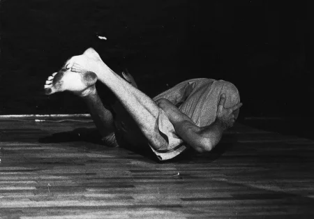 Aktor ubrany w jasne spodenki. Jest boso i ma nagi tors. Wygląda, jakby miał się przeturlać na podłodze.