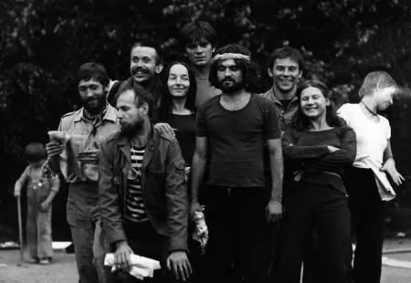 Plener. Grupa aktorów i aktorek pozuje do zdjęcia. Stoją bardzo blisko siebie, uśmiechają się. Wszyscy mają ciemne ubrania poza wyróżniającą się aktorką z prawej strony. Aktorka ubrana jest w białą podkoszulkę i patrzy w bok. Na dalszym planie po lewej stronie stoi dziecko bawiące się kijem. W tle widać drzewa.