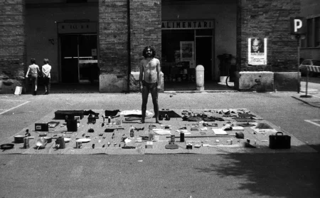 Przestrzeń miejska. Przy ulicy leży rozłożona płachta, na której leżą przedmioty, między innymi kurtki, koszulki, buty, butelki, przybory szkolne. Na płachcie stoi zwrócony twarzą do widza aktor. Ubrany jest tylko w majtki. Za jego plecami widać budynek i przechodniów.