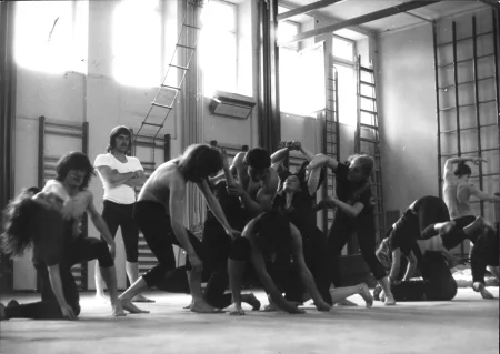 Grupa aktorów i aktorek na sali gimnastycznej. Wykonują wspólne akrobacje, są w bliskim kontakcie fizycznym. Niektórzy ubrani są w czarne spodenki i czarne podkoszulki, inni mają nagie torsy. Wszyscy są boso. W pewnej odległości od grupy stoi aktor ubrany w czarne spodenki i białą podkoszulkę. Ma długie włosy i wąsy. Patrzy na grupę, jakby był trenerem.