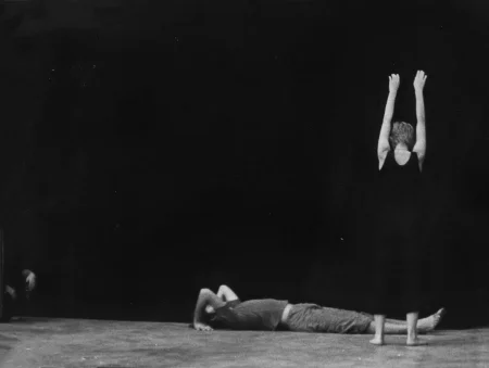 Po prawej stronie, odwrócona tyłem do widza, stoi aktorka. Ubrana jest na czarno, jest bosa. Obie ręce trzyma wyciągnięte ku górze. Z tyłu sceny, przed aktorką, leży aktor. Trzyma obie ręce za głową. Ubrany jest w ciemne spodnie i ciemną podkoszulkę. Jest boso.