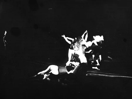 Niewyraźne, prześwietlone zdjęcie. Z przodu sceny troje aktorów siedzi lub klęczy na podłodze. W głębi sceny grupa aktorów stoi w ekspresyjnych pozach. Po lewej stronie z cienia wyłania się ledwo widoczna psotać.