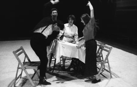 Przy stole stoi pięcioosobowa grupa aktorów i aktorek. Wszyscy wyglądają na zdenerwowanych. Niektórzy unoszą stół, inni w niego uderzają.