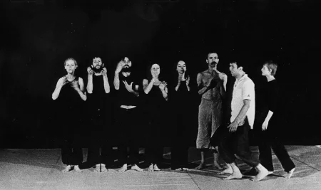 Grupa aktorów i aktorek stojących w szeregu na tle czarnej płachty. Wszyscy są boso i ubrani w czarne ubrania. Wyróżnia się aktor ubrany w jasne spodenki. Z prawej strony idzie grupa aktorów i aktorek zwróconych profilem w stronę widza.