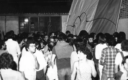 Przestrzeń miejska. Tłum ludzi. W tle stoi budynek z wielkimi oknami. Z prawej strony stoi płachta z niewyraźnym rysunkiem.