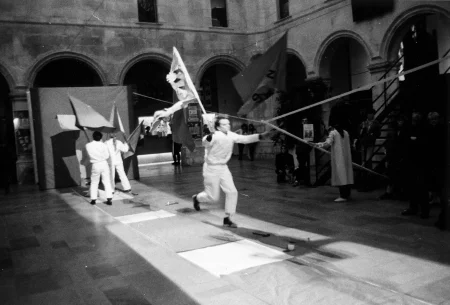 Plac wewnątrz budynku. Na podłodze leży rozwinięty wybieg. Na końcu wybiegu, pod ścianą stoi grupa aktorów i aktorek odwróconych do widza plecami. W rękach trzymają flagi. Wzdłuż wybiegu biegnie aktor trzymający w ręce flagę. Wszyscy aktorzy ubrani są na biało. Obok wybiegu stoi niewielka grupa widzów. W tle widać kolumny budynku.