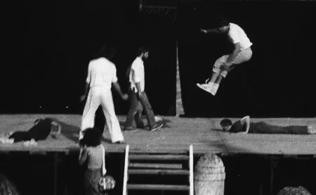 Scena plenerowa. Na scenę prowadzą drewniane schody. Z tyłu sceny ściana z czarnej płachty. Pod sceną stoi widzka. Na scenie, po lewej stronie na plecach leży aktorka ubrana na czarno. Obok niej stoi zwrócony plecami do widza aktor ubrany na biało. Obok niego przechodzi zwrócony profilem do widza aktor ubrany w jeansy i białą koszulę. Z prawej strony sceny bardzo wysoko skacze aktor. Ubrany jest w jasne buty sportowe, jasne spodnie i jasną podkoszulkę. Za nim na brzuchu leży aktor ubrany na czarno.