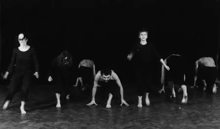 Grupa aktorów i aktorek wygląda, jakby przygotowywała się do biegu. Jeden aktor zwrócony jest twarzą w stronę widza, reszta zwrócona jest do widza plecami. Wyróżniają się trzy aktorki, które stoją. Są zwrócone twarzą w stronę widza. Ubrane są na czarno.