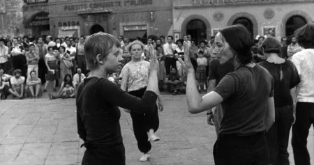 Performance uliczny. Na pierwszym planie stoją dwie aktorki zwrócone profilem do widza i twarzą do siebie. Pokazują sobie wnętrza dłoni. Z tyłu, w centrum sceny idzie przed siebie zwrócona twarzą do widza aktorka. Ubrana jest w czarne spodnie i jasną koszulkę. Z prawej strony stoi słabo widoczna grupa aktorów i aktorek. W tle stoi i siedzi duża grupa widzów. Za ich plecami stoją budynki.