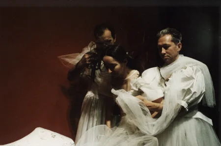 W środku między dwoma aktorami stoi aktorka. Wszyscy mają na sobie białe suknie ślubne i patrzą w dół. Aktor z lewej strony dotyka włosów aktorki. Z tyłu widać czerwoną ścianę.