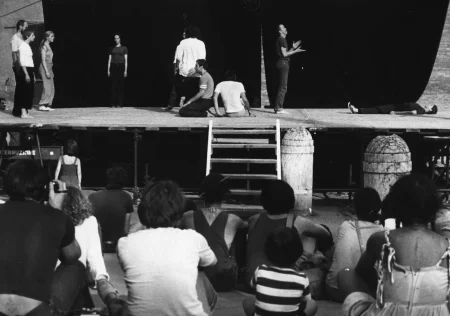 Scena plenerowa. Na scenę prowadzą drewniane schody. Z tyłu sceny ściana z czarnej płachty. Pod sceną siedzi grupa widzów. Na scenie, po lewej stronie, grupa aktorów i aktorek zwróconych profilem do widza. Stoją w miejscu. Obok nich stoi aktorka zwrócona twarzą do widza. Ubrana jest w czarne spodnie i czarną podkoszulkę. W centrum sceny dwaj aktorzy stoją zwróceni do siebie twarzami. Tuż rzy nich dwóch aktorów siedzi na scenie. Obok nich stoi zwrócony profilem do widza aktor ubrany na czarno. Aktor wygląda, jakby klaskał. Po prawej stronie sceny leży brzuchem do góry aktorka. Ubrana jest na czarno.