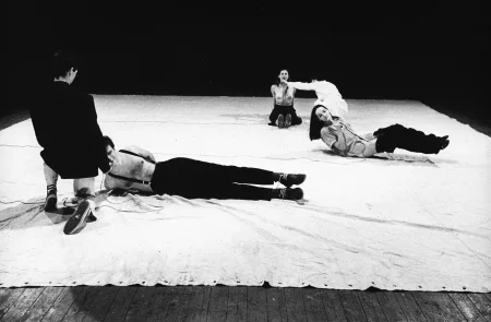 Pięcioosobowa grupa aktorów i aktorek na białej płachcie. Z przodu klęczy aktor zwrócony plecami w stronę widza. Trzyma mikrofon przy aktorze, który leży zwrócony twarzą w stronę widza. Za nimi leży aktorka z mikrofonem przy ustach. Z tyłu kuca aktorka zwrócona plecami w stronę widza. Trzyma mikrofon przy aktorce kucającej twarzą w stronę widza.