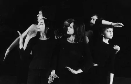 Dwóch aktorów i cztery aktorki. Aktor stojący po lewej stronie nie ma koszulki, jest poważny i trzyma dłoń przy swoim czole. Aktorki ubrane są na czarno i wykonują gesty dłońmi. Stojący za nimi aktor jest ledwo widoczny, ale widać, że patrzy przed siebie.