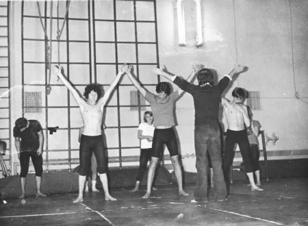 Grupa aktorów i aktorek na sali gimnastycznej. Z tyłu, pod ścianą stoi drabina gimnastyczna. Aktorzy i aktorki zwróceni przodem do widza wyglądają, jakby ćwiczyli. Wszyscy są boso, ubrani w strój sportowy. Dwóch aktorów ma nagi tors. Przed grupą, tyłem do widza stoi aktor z uniesionymi rękami. Wygląda, jakby był trenerem grupy.