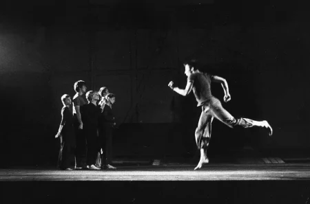 Po lewej stronie pustej sceny stoi zwarta grupa aktorów i aktorek. Są boso, mają na sobie czarne ubrania. Patrzą na aktora po prawej stronie. Aktor biegnie lewą stronę. Jest bosy i ubrany w ciemne ubrania. Przestrzeń wygląda na industrialną.