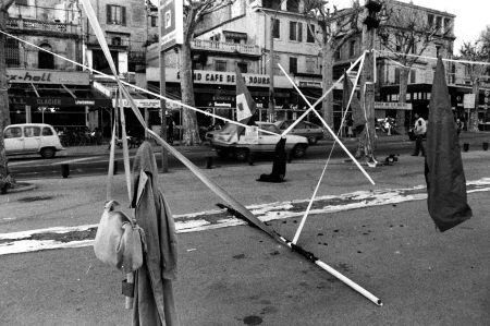 Przestrzeń miejska. Na środku ulicy rozwinięty wybieg. Nad wybiegiem wisi dużo linek, na których wiszą ubrania i fragmenty materiałów. Wybieg jest brudny. W tle widać budynki, samochody i przechodniów.
