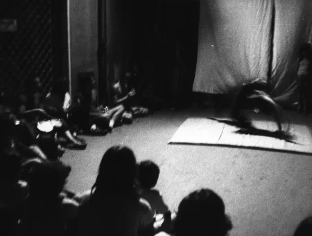 Niewyraźne zdjęcie. Przy scenie siedzi zgromadzona widownia. Na scenie leży mata, na której aktor wykonuje akrobacji. Z tyłu widać białą kurtynę. Pod kurtyną stoi aktorka zwrócona twarzą w stronę widza.