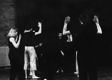 Aktorzy i aktorki stoją zwróceni plecami w stronę widza. Mają lekko uniesione ręce. W tle widać aktora zwróconego twarzą w stronę widza. Aktor się uśmiecha. Po lewej stronie widać ceglaną ścianę i biały kaloryfer.