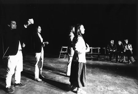 Przestrzeń miejska. Grupa aktorów i aktorek rozwija na ulicy między budynkami wielką biała płachtę. Po lewej stronie przechodzi aktorka z wiadrem i szczotką.