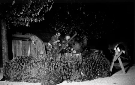 Grupa aktorów przebranych za żołnierzy stoi w okopie przodem w stronę widza. Wyglądają, jakby strzelali z karabinów. W tle widać drzewa i budynek z blachy.