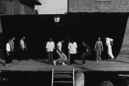 Scena plenerowa. Na scenę prowadzą drewniane schody. Z tyłu ściana z czarnej płachty. Pośrodku, zza płachty wystaje latarnia. Na scenie stoi grupa aktorów i aktorek. Aktorzy i aktorki po lewej stronie zwróceni są profilem do widza. Wyglądają, jakby powoli szli przed siebie. W centrum dwóch aktorów zwróconych w stronę widza idzie do przodu sceny. Ubrani są w jeansy i białe koszule. Obok nich stoi dwóch aktorów zwróconych profilem do widza i twarzami do siebie nawzajem. Po prawej stronie stoją aktorka i aktor. Oboje ubrani są w kombinezony - aktorka w jeansowy, aktor w biały. Za sceną widać budynki.