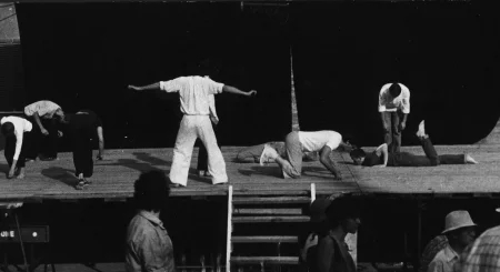 Scena plenerowa. Na scenę prowadzą drewniane schody. Z tyłu sceny ściana z czarnej płachty. Pod sceną przechodzi grupa widzów. Na scenie, po lewej stronie troje aktorów. Stoją nisko pochyleni ku scenie. Obok nich stoi zwrócony plecami do widza aktor ubrany na biało. Aktor ma ręce wyciągnięte do boków. Po prawej stronie dwóch aktorów zwróconych do siebie twarzami kładzie się na scenę brzuchem do dołu. Za nimi stoi lekko pochylony aktor z głową zwieszoną w dół. Ubrany jest w jeansy i białą koszulę.