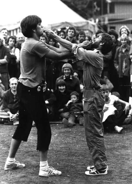 Dwóch aktorów stoi naprzeciwko siebie. Są ubrani w trampki, spodnie i podkoszulki. Podpalają sobie wzajemnie papierosy, które trzymają w ustach. Na drugim planie widać grupę widzów oglądających aktorów.