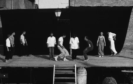 Scena plenerowa. Na scenę prowadzą drewniane schody. Z tyłu sceny ściana z czarnej płachty. Pośrodku, zza płachty wystaje latarnia. Na scenie stoją aktorzy i aktorki w różnych działaniach. Aktorzy i aktorki po bokach sceny są statyczni. Aktorzy w centrum sceny dynamicznie się ruszają. Wyglądają, jakby mieli na sobie prywatne ubrania. Wspólnie tworzą choreograficzną kompozycją.