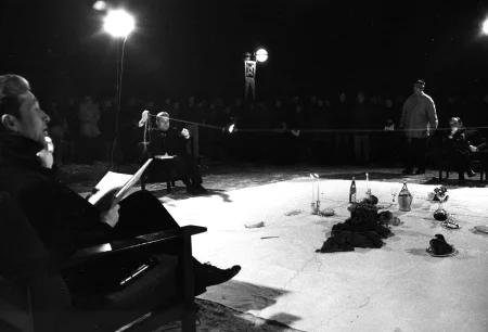 Na białej płachcie leżą przedmioty, takie jak kieliszki, świece, owoce i butelki. Po lewej stronie płachty na krześle siedzi aktor. Ubrany jest na czarno. W rękach trzyma książkę i mówi coś do aktora siedzącego na krześla po drugiej stronie płachty. Z tyłu płachty na krześle siedzi aktor trzymający w rękach napiętą linkę. Za jego plecami stoi reflektor i grupa widzów.