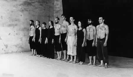 Grupa aktorów i aktorek stojących w szeregu na tle czarnej płachty. Aktorzy ubrani są w czarne spodenki, mają nagie torsy. Aktorki ubrane są w czarne spodnie i czarne koszulki. Wśród grupy wyróżnia się aktor ubrany w jasne spodenki. Wszyscy są boso. Wyglądają, jakby śpiewali.